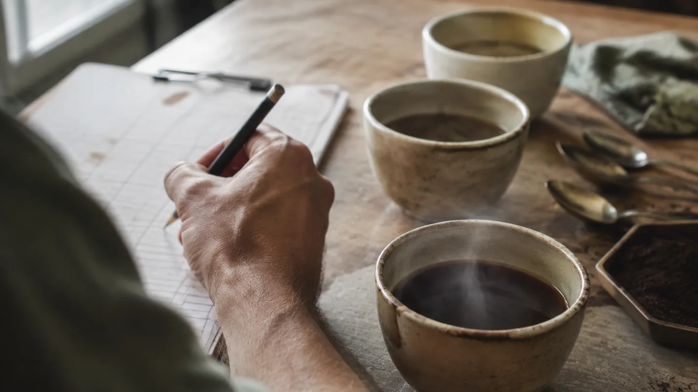 A brewer notes changes as three cups cool on a worn tasting table.