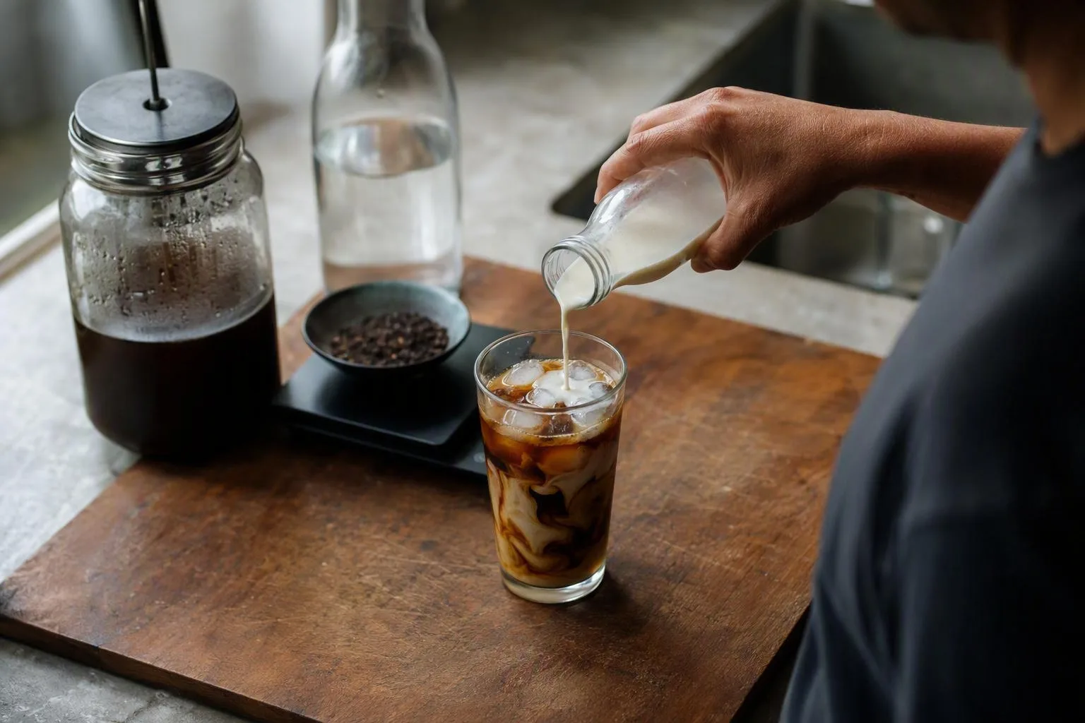 A hand pours milk into an iced cold brew beside the measured ingredients for the batch.