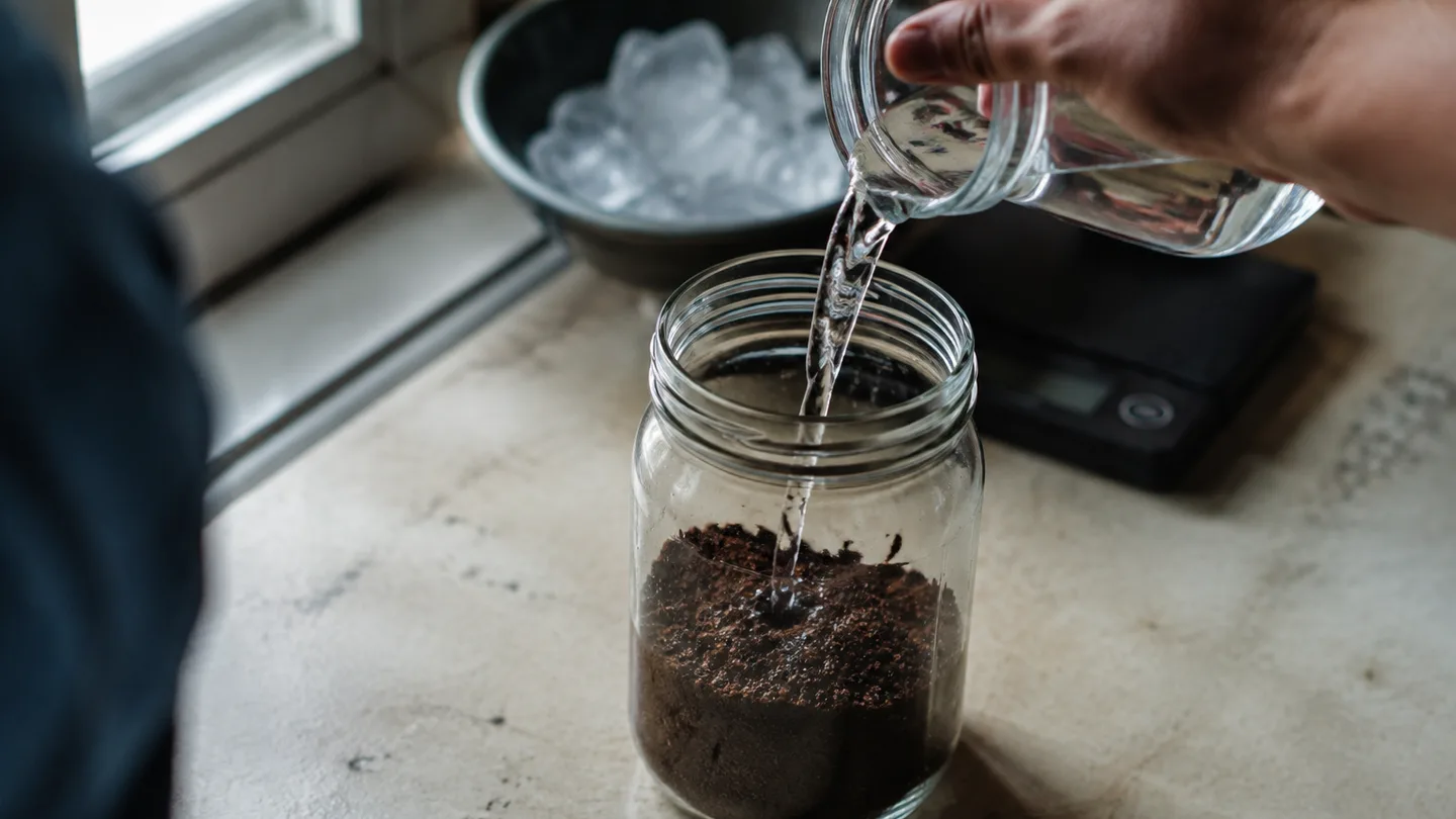 A hand pours water over coarse grounds in a glass jar before the long steep.