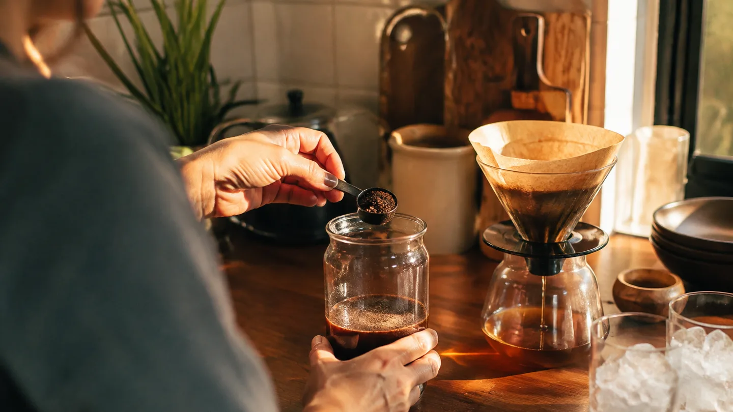 A hand measures coarse grounds into a glass jar beside a slow filter, showing the careful brewing process.