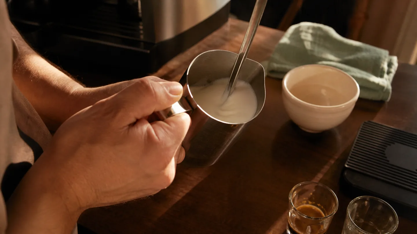A hand folds glossy milk foam in a steel pitcher beside an espresso cup, showing a smooth, pourable texture.