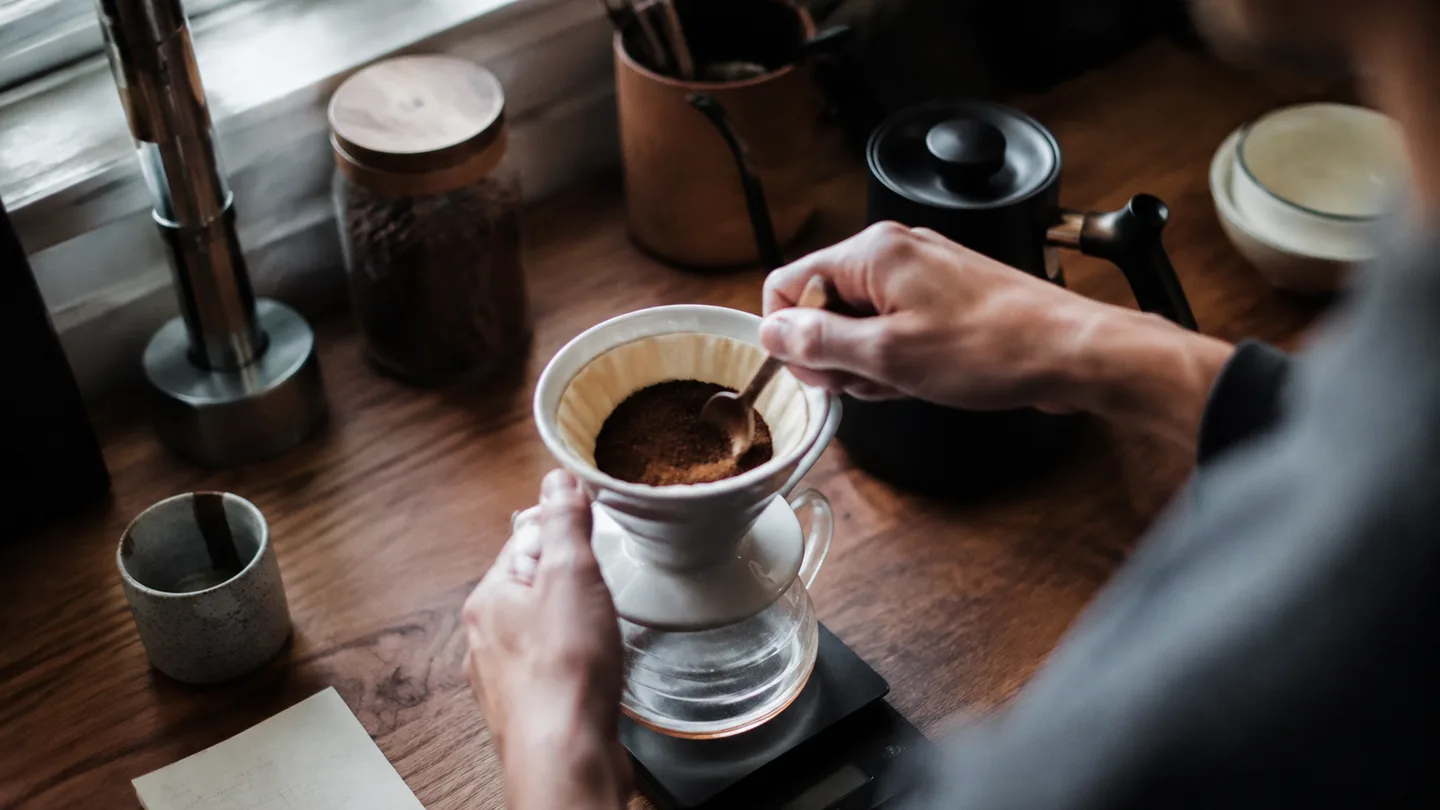 Hands weigh coffee grounds beside a dripper and carafe, establishing a careful repeatable morning brewing routine.