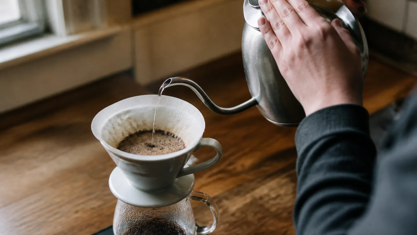 A kettle sends a small stream onto coffee grounds as the bloom wets the bed evenly.