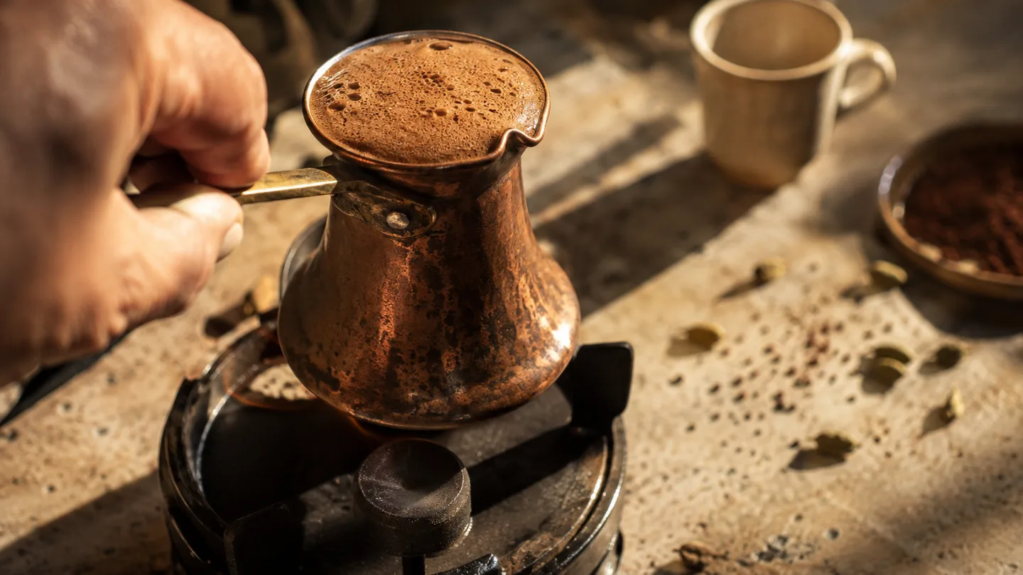 Foam swells to the rim of a copper cezve as a hand lifts it from the heat.