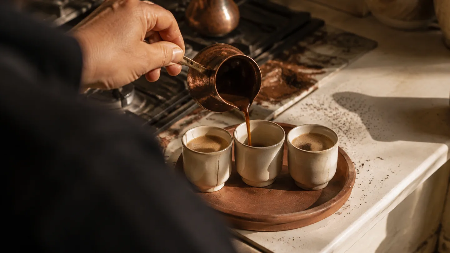 A hand pours foamy coffee into one of three small cups to keep a shared batch even.