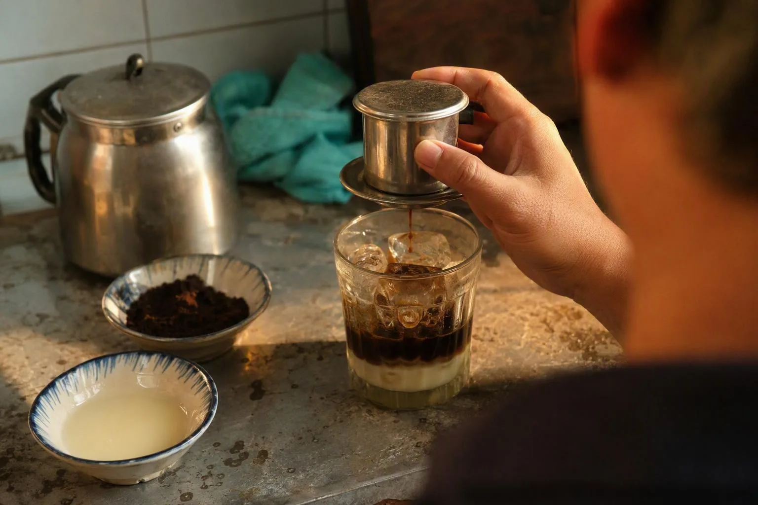 A phin drips coffee over ice and condensed milk, with measured ingredients ready on a worn counter.