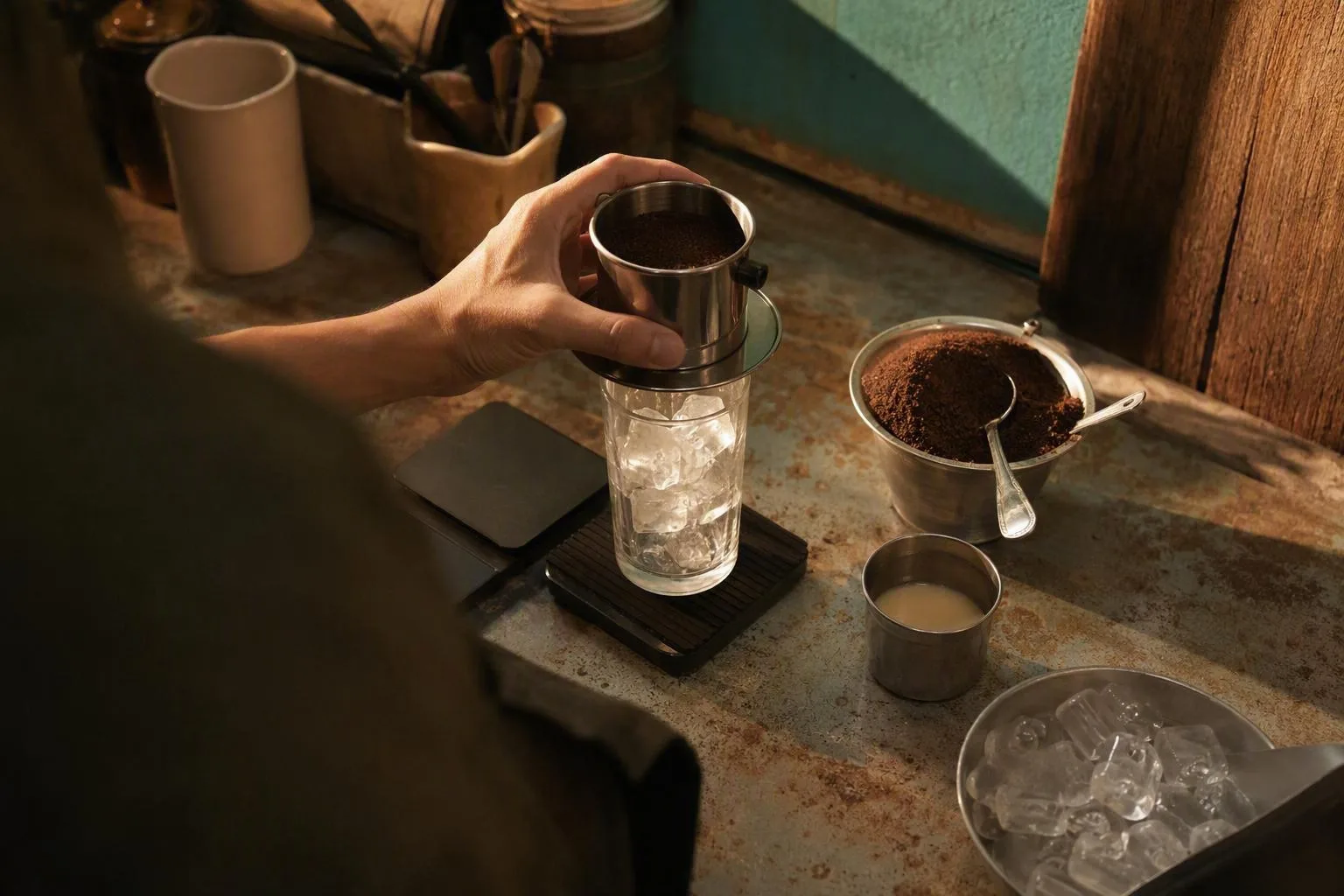 A phin, coffee grounds, condensed milk, and ice are measured on a worn counter before brewing.