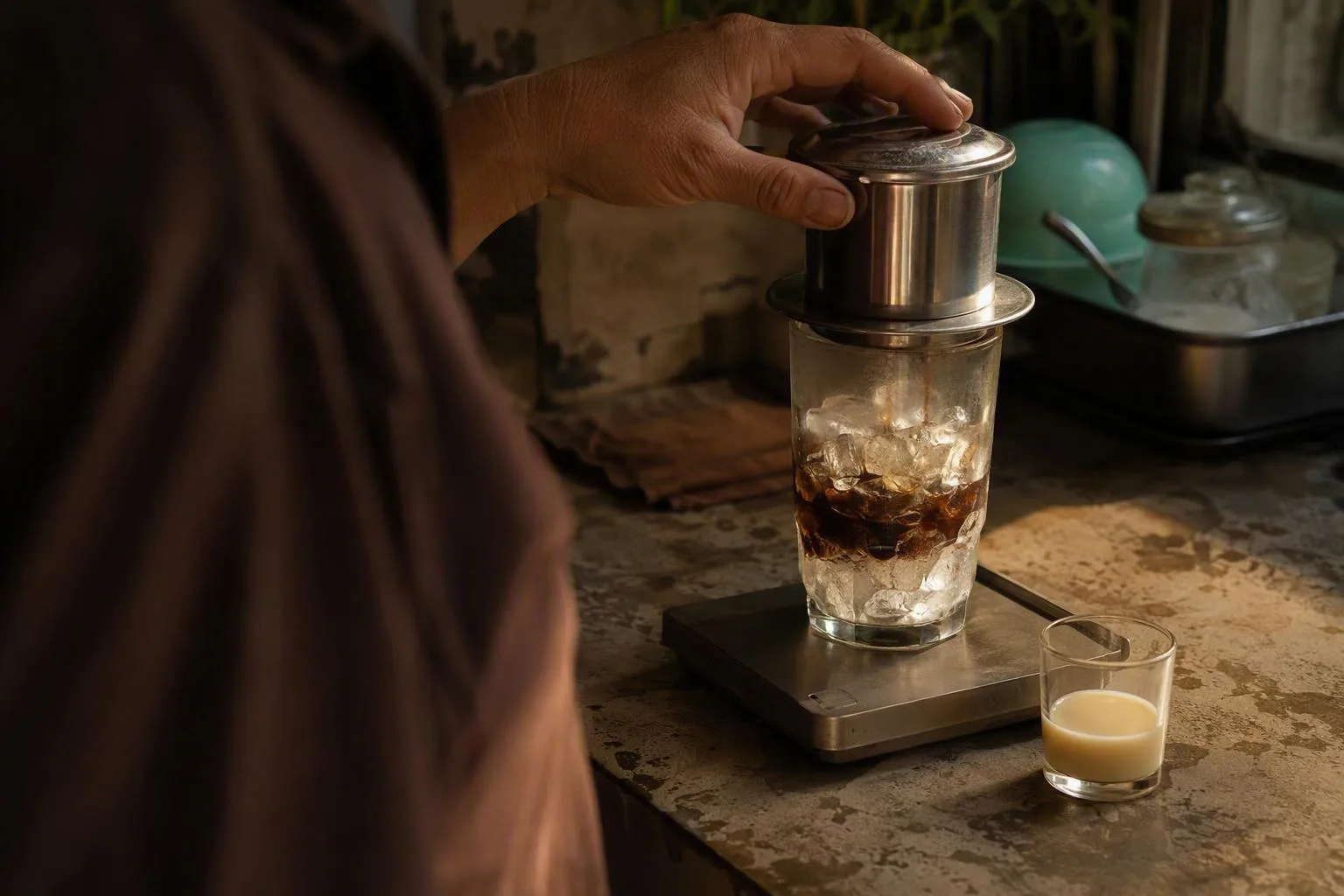 A hand pours hot coffee from a phin over ice on a scale beside milk, showing the brew being diluted.