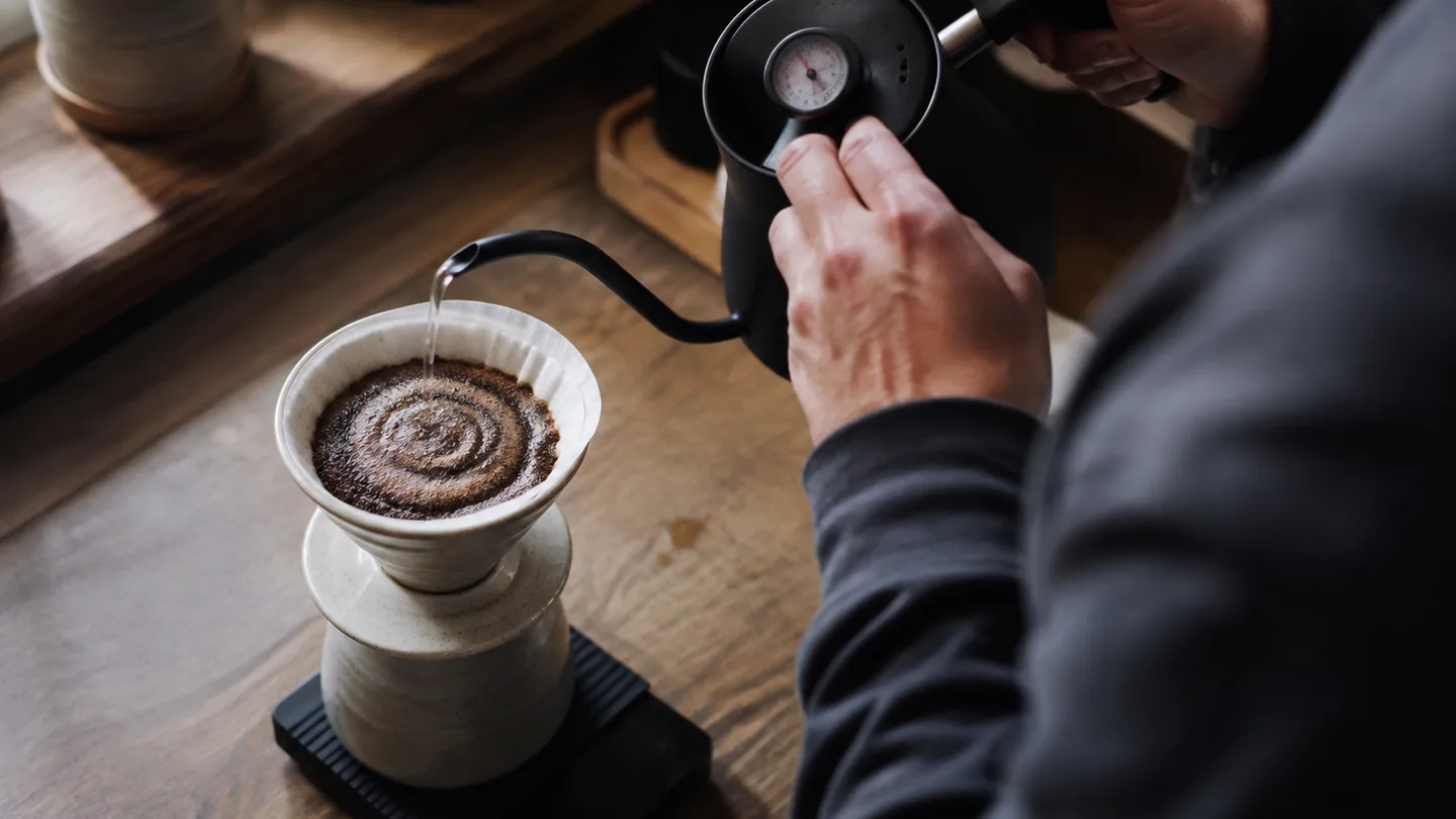 A hand pours hot water over coffee grounds beside a thermometer and scale, showing a measured brewing start.