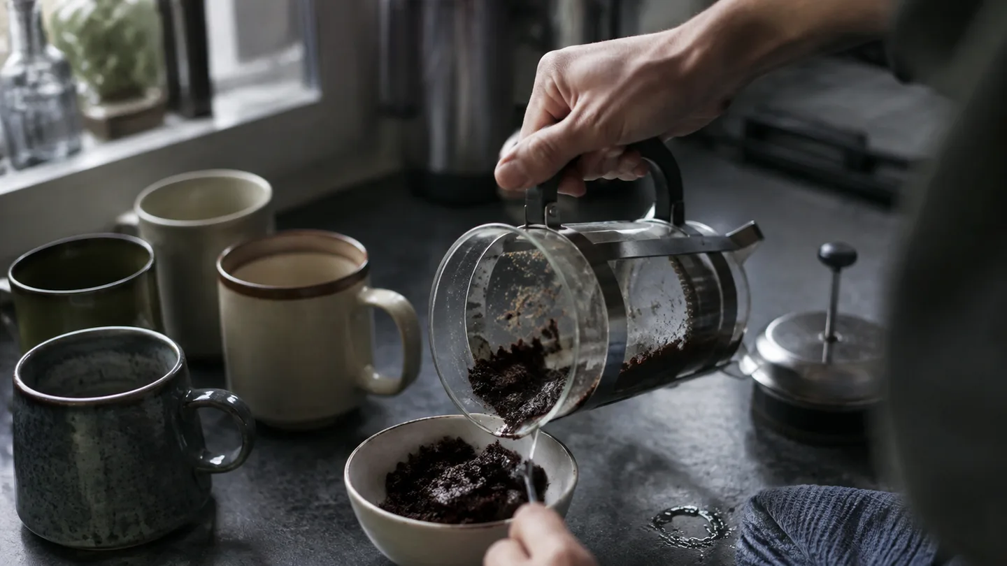 A hand scrapes wet coffee grounds from a used press beside waiting mugs, showing the cleanup after serving.