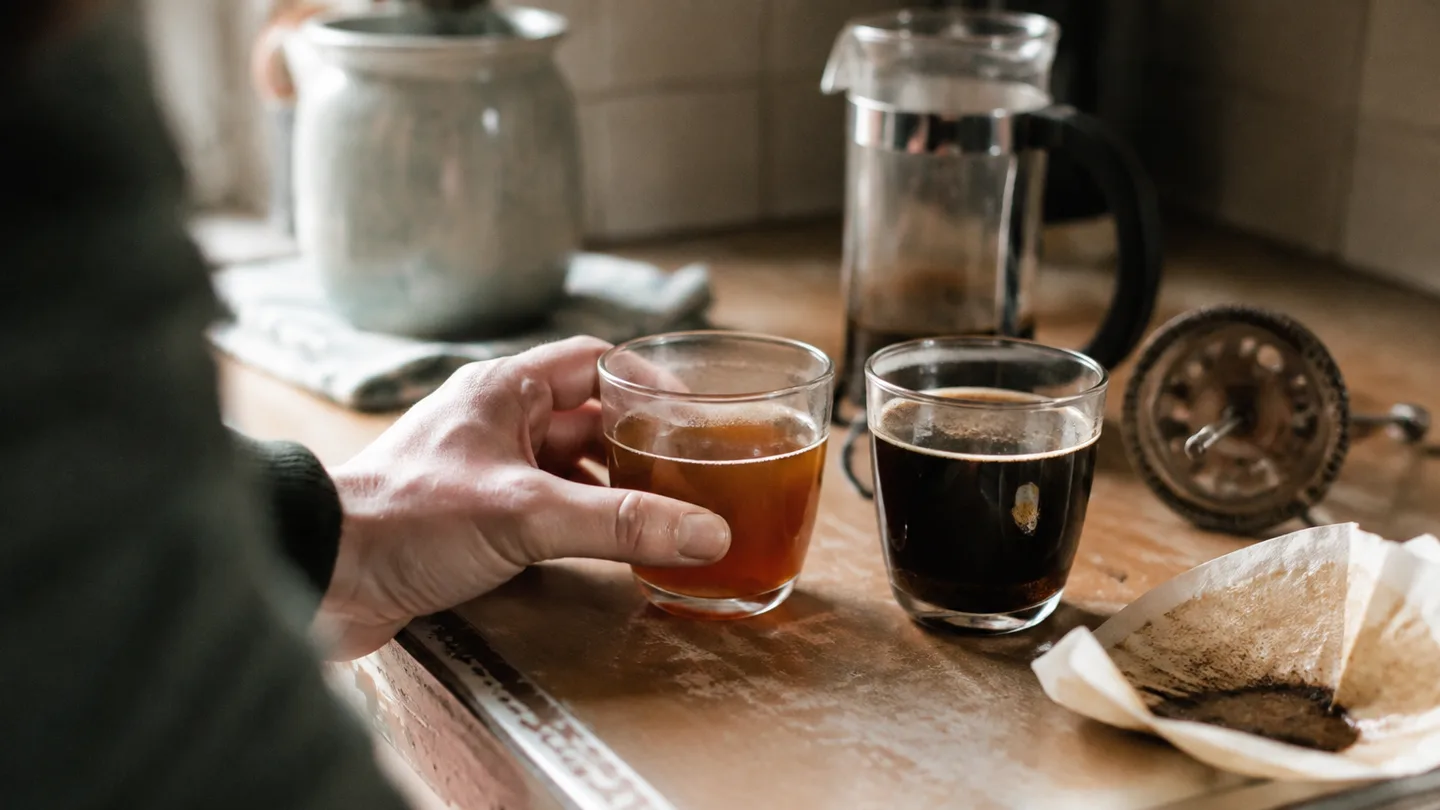 A hand compares a clear filtered brew with a darker cup showing oil sheen and settled fine sediment.